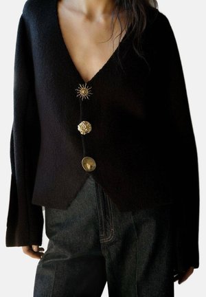Femme portant un pull noir à col en V avec trois boutons dorés uniques et un pantalon sombre taille haute, debout avec les bras relâchés.