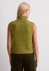 Mouwloze, olijfgroene gebreide top met een hoge ribbelkraag. Textuurstof met een losse pasvorm, gecombineerd met een bruine corduroy broek.