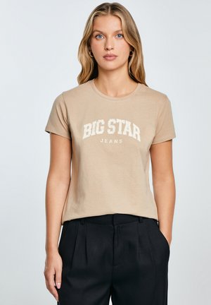Béžové bavlněné tričko s nápisem "BIG STAR JEANS" bílým písmem, kulatým výstřihem a krátkými rukávy, v kombinaci s černými skládacími kalhotami.