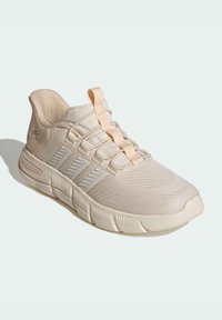 Zapatilla deportiva beige con parte superior de malla texturizada, detalles en rosa claro y tres franjas blancas en el lateral. Punta redondeada y suela acolchada.