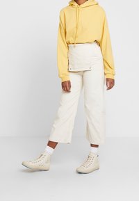 Felpa gialla con coulisse, pantaloni cropped in corduroy bianchi e sneakers alte beige. L'outfit presenta una tasca frontale sui pantaloni.