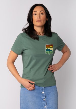 T-shirt en coton vert avec un col rond, ornée d'un graphique coloré sur la poitrine gauche représentant des montagnes, un soleil, et le texte "GOOD VIBE".