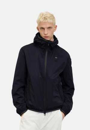 Giubbotto leggero blu navy con chiusura a cerniera frontale, cappuccio regolabile e tasche laterali. Presenta polsini elasticizzati e un design minimalista.