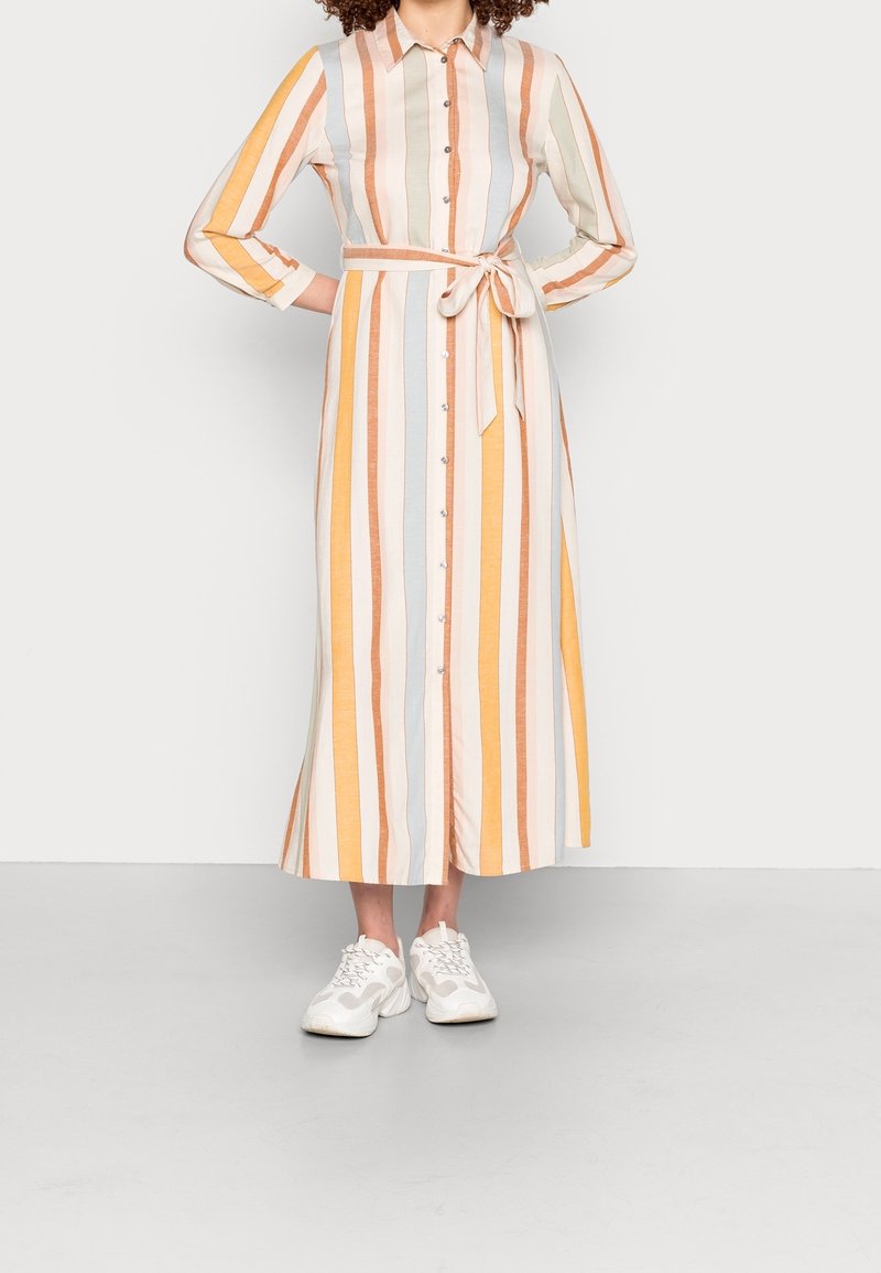 Femme portant une longue robe boutonnée à rayures verticales pastel jaune, orange et bleu, associée à des baskets blanches.