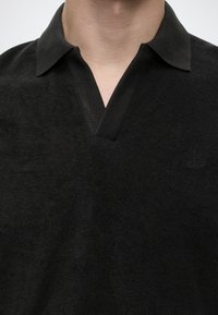 Personne portant une chemise noire à col avec une ouverture en V, laissant apparaître une partie du cou et du haut de la poitrine.