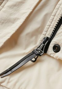 Beżowa tkanina o teksturowanym wykończeniu, z czarnym zamkiem błyskawicznym oraz akcentami w kolorze czarnym. Zawiera czarną linkę do regulacji.