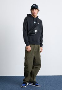 Crna dukserica s plavim Nike logotipom, maslinasto zelene cargo hlače, crna fanny pack torba s bijelim logotipom, plavo-crne tenisice i crna kapica.