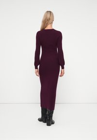 Langes, eng anliegendes Kleid aus tiefviolettem Strickstoff, mit langen Ärmeln und Rundhalsausschnitt, kombiniert mit schwarzen, hohen Stiefeletten.