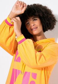 Felpa gialla brillante con maniche oversize, dettagli rosa e testo grafico audace. Presenta un cappuccio con lacci e una texture liscia.
