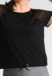 Svart sporttröja med genomskinliga meshärmar, rundad halsringning och justerbar dragsko i nederkant för passform. Mjuk tyg med avslappnad design.