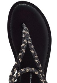 Sandalia tipo thong negra con correas trenzadas adornadas con pequeños acentos metálicos. Suela negra suave con plantilla acolchada.