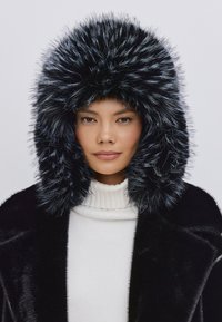 Faux fur hætte i sort med sølvspidser, indrammet af en blød, skinnende sort frakke. Høj krave hvid sweater nedenunder. Glat, struktureret overflade.