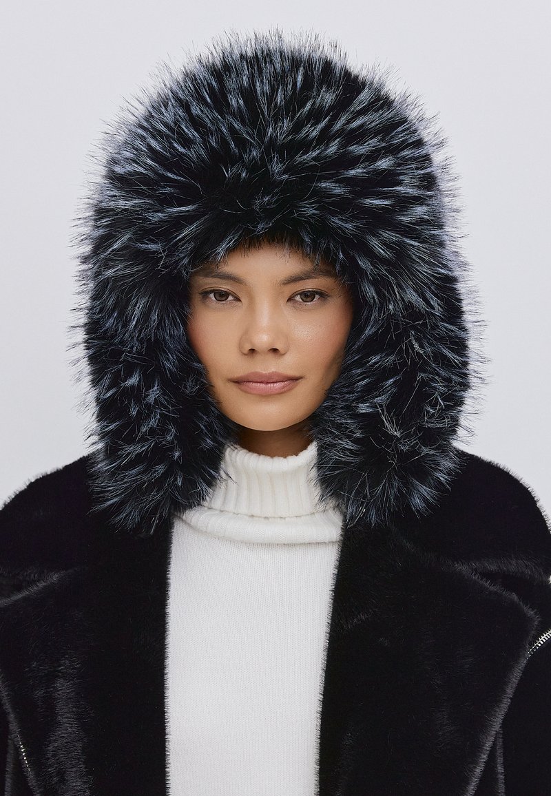 Faux fur hætte i sort med sølvspidser, indrammet af en blød, skinnende sort frakke. Høj krave hvid sweater nedenunder. Glat, struktureret overflade.
