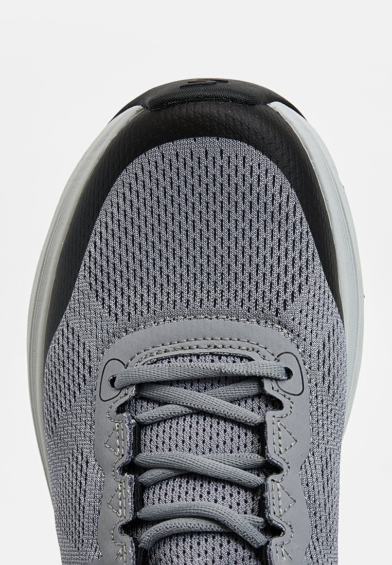 Zapatilla deportiva gris con parte superior de malla, cordones redondos y acentos de goma negra. La suela es gruesa y ligeramente curvada. Acabado texturizado.