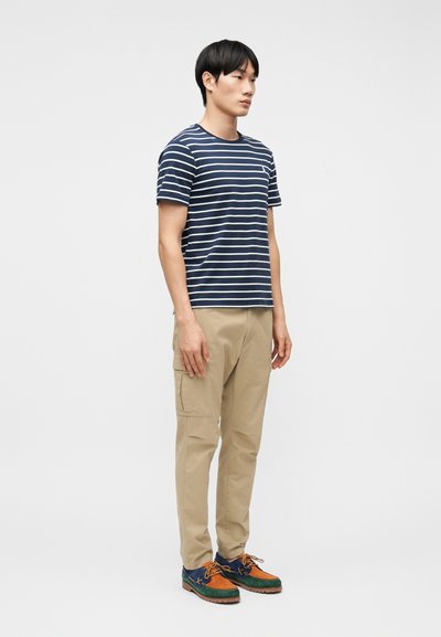 Νεαρός άνδρας που φοράει μπλουζάκι με ναυτικές και λευκές ρίγες, μπεζ παντελόνι καμπαρντίνα και πολύχρωμα καθημερινά παπούτσια, στέκεται μπροστά σε ένα απλό φόντο.