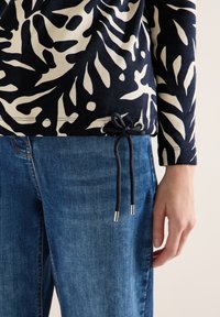 Person trägt blaue Jeans und ein dunkelblau-weißes, abstrakt gemustertes Langarmshirt mit schwarzer Kordel, die an der Taille gebunden ist.