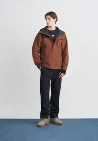 Καφέ αδιάβροχο μπουφάν με μαύρες λεπτομέρειες, κλείσιμο με φερμουάρ και κουμπιά, μπροστινές τσέπες, μαύροι παντελόνια καμπαρντίνας και μπεζ μπότες. Απλός και λειτουργικός σχεδιασμός.