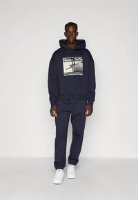 Hooded sweatshirt and matching sweatpants in dark navy, featuring a graphic print on the front. White sneakers complete the outfit.