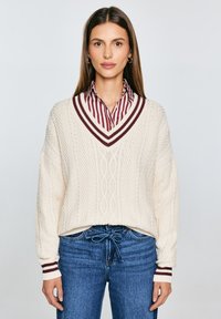Elfenbeinfarbener Strickpullover mit V-Ausschnitt und Kabelmuster; verfügt über bordeauxfarbene und weiße gestreifte Akzente am Kragen und den Manschetten. Kombiniert mit blauen Jeans.