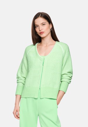 Young woman with long brown hair wearing light green textured zip-up sweater and matching pants standing with one hand in pocket.