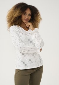 Camisa blanca de manga larga con tejido de ojal, que presenta un escote en V y mangas voluminosas. Combinada con pantalones ajustados verde oliva.