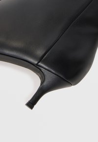Chaussure à talon haut en cuir noir avec une texture lisse, bout pointu et petit talon carré. Le design présente des détails de couture minimalistes.