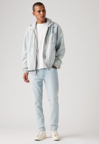 Lyseblå denimjakke med grå hette, brukt over en hvit t-skjorte, kombinert med lyseblå jeans og hvite sneakers.