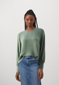 Blusa de manga larga en un verde claro con un acabado texturizado, escote redondo y corte holgado, combinada con una falda de mezclilla azul.