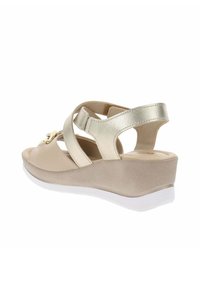 Sandali con zeppa beige con una fascia metallica dorata, dotati di una finitura liscia e chiusura regolabile. La suola bianca offre un contrasto.