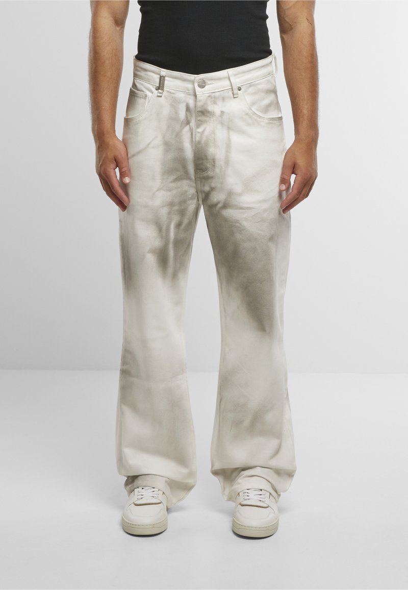 Homme portant un pantalon large blanc, ample, légèrement taché, avec des baskets blanches et un haut noir sans manches, sur un fond uni.