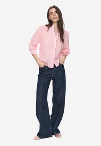 Lichtroze blouse met oogjesstof, lange mouwen, knoopsluiting aan de voorkant en geplooide schouders, gecombineerd met donker denim jeans met wijde pijpen en roze ballerina's.