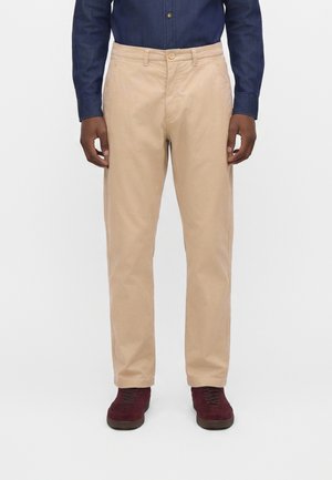Hombre que lleva pantalones beige de corte recto, camisa azul oscuro abotonada y zapatos granate con cordones, de pie frente a un fondo blanco liso.
