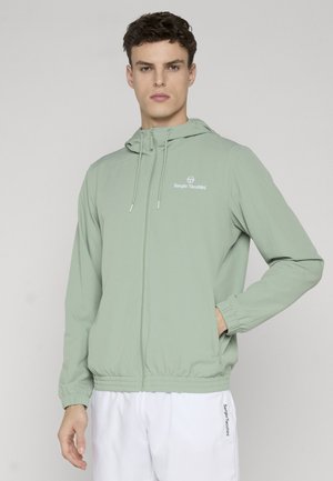 Ung mann iført en lysegrønn hettejakke med glidelås og hvite shorts med merkevarelogoer, poserer med den ene hånden i lommen mot en ensfarget bakgrunn.