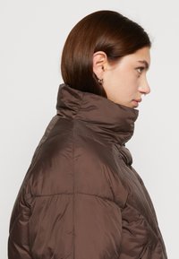 Veste matelassée marron avec un col haut, texture lisse et segments cousus, vue de côté. Présente un design épuré et un matériel minimal.