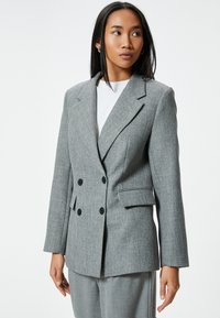 Blazer gris de corte doble en tejido texturizado. Presenta solapas con muesca, dos bolsillos frontales y botones negros. Ajuste entallado con hombros estructurados.