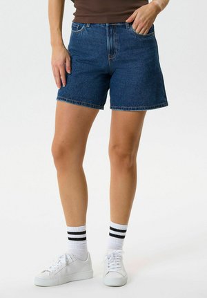 Inimene, kes kannab siniseid teksapükse, valgeid tossusid ja valgeid sokke kahe musta triibuga, seisab ühe käega taskus.