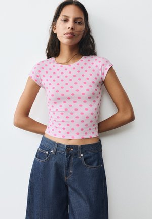 Ung kvinde med langt mørkt hår iført en lyserød t-shirt med prikker og højtaljede blå jeans, stående mod en ensfarvet hvid baggrund.