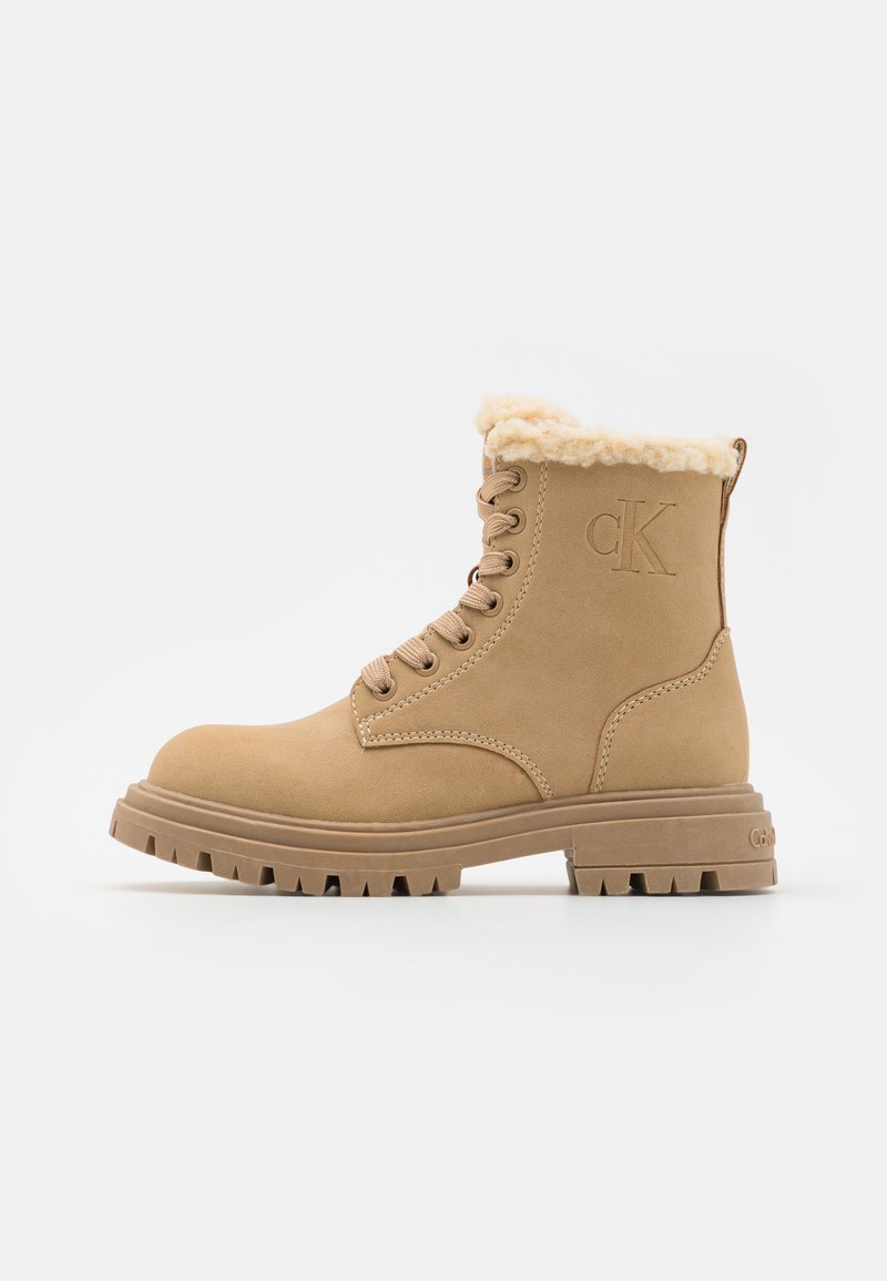 Tan ankle boot with faux fur lining, round toe, padded collar, and lace-up front. Features textured rubber outsole and embossed logo detail.