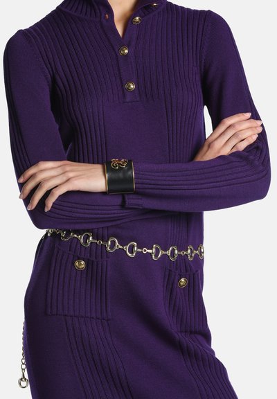 Femme portant une robe violet côtelée avec des boutons dorés, une ceinture chaîne dorée, les bras croisés et un bracelet manchette noir avec un détail doré.