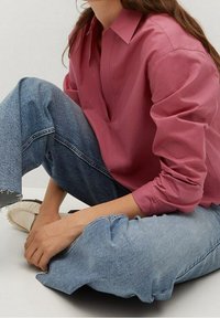 Chemise rose à manches longues avec col, présentant un décolleté en V ; associée à un jean bleu clair à coupe décontractée avec des ourlets effilochés.