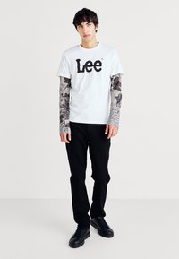 Hvit t-skjorte med svart "Lee"-logo, lagdelt med mønstrede grå langermede skjorter. Svart bukser og svarte sneakers fullfører antrekket.