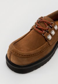 Chaussure en daim marron avec un bout rond, ornée de lacets rouges et d'accents métalliques argentés. La semelle est noire et rainurée pour une meilleure adhérence.