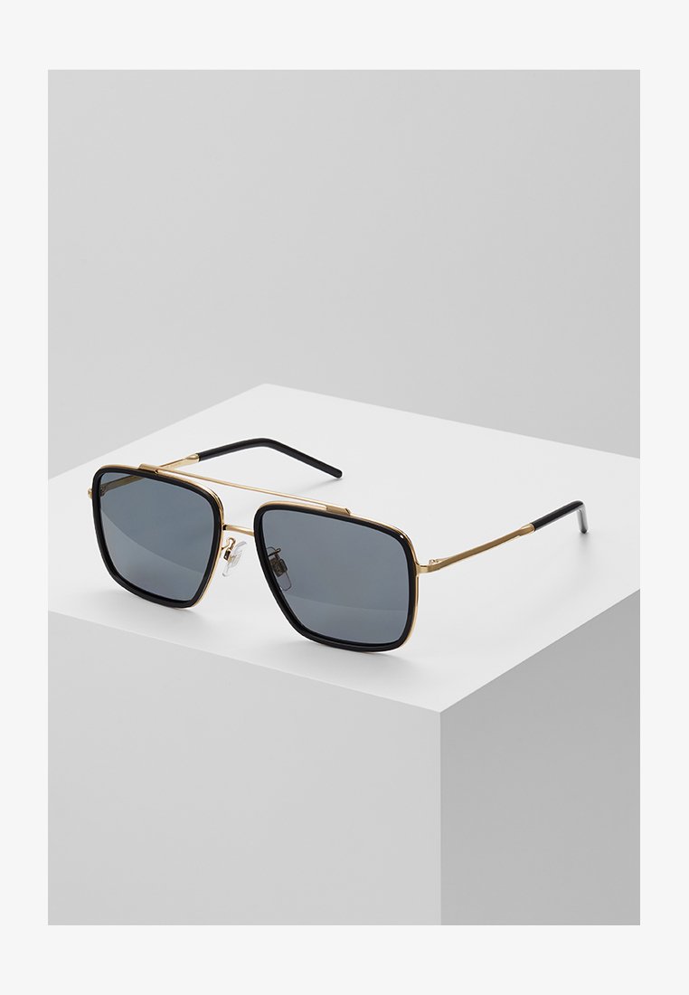 Óculos de sol com um design quadrado, armações pretas, detalhes em ouro, lentes escuras e hastes de metal, expostos sobre uma superfície cinza-clara.