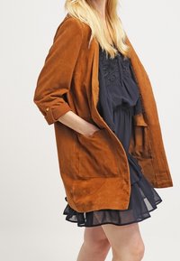 Brun mocka oversized jacka med avslappnad passform, uppvikt ärm, och framfickor, lager på lager  över en mörk klänning. Slät textur.