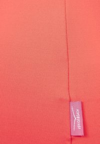 Tejido de coral brillante con una textura acanalada y una pequeña etiqueta cosida en rosa, ubicada cerca del borde.