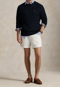 Navy cable-knit sweater, white shorts, leather loafers. Sweater features a small red logo. Layered with a blue striped collared shirt.