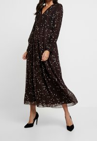 Lungo vestito marrone scuro con scollatura a V, maniche lunghe e motivi a foglia bianchi. Tessuto leggero con una gonna fluttuante, abbinato a tacchi neri.
