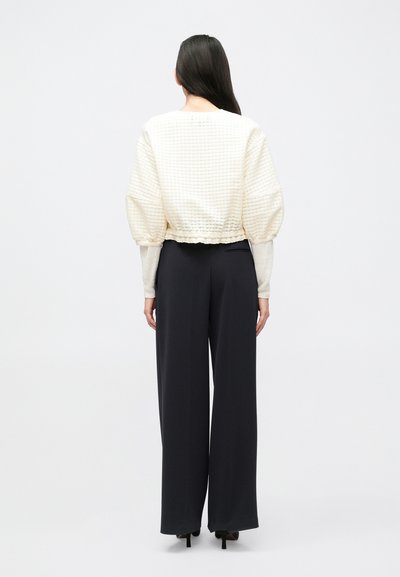 Μπλούζα με μανίκια φουσκωτά σαν κρέμα γάλακτος, κατασκευασμένη από υφασμάτινη υφή, συνδυασμένη με μαύρο παντελόνι ψηλόμεσο με φαρδιά γραμμή και ουδέτερα παπούτσια.