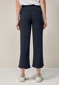Pantalons larges bleu marine avec une texture lisse, une taille élastique et deux poches arrière. Caractéristiques des ourlets arrondis et assortis avec des baskets blanches.