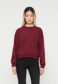 Ung kvinna med långt ombre hår som bär en vinröd tröja med broderade ärmar och svarta byxor, stående framför en enkel bakgrund.